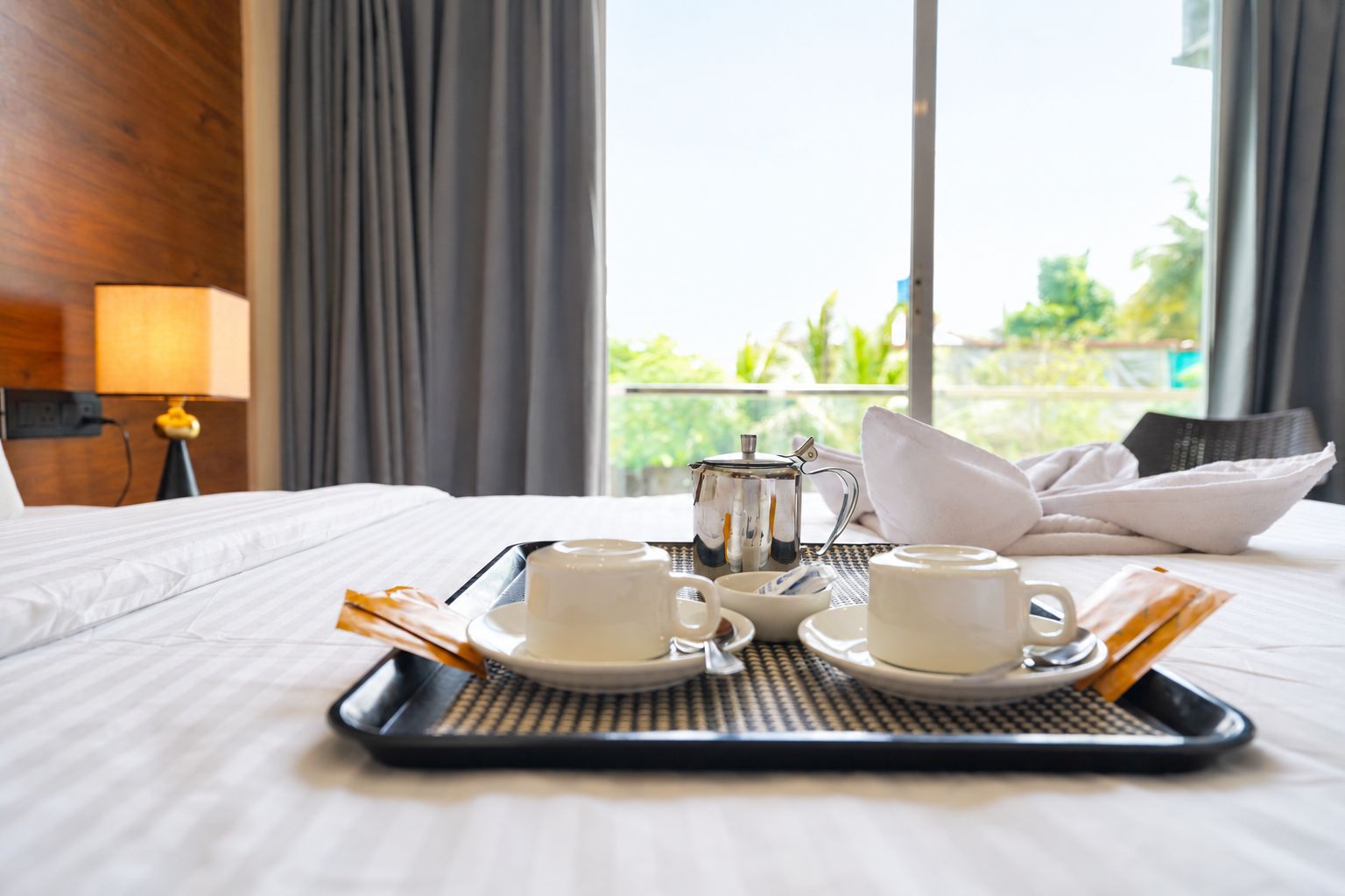 A hotel room with a bed, a tray with two cups and a teapot, and a view of greenery outside.