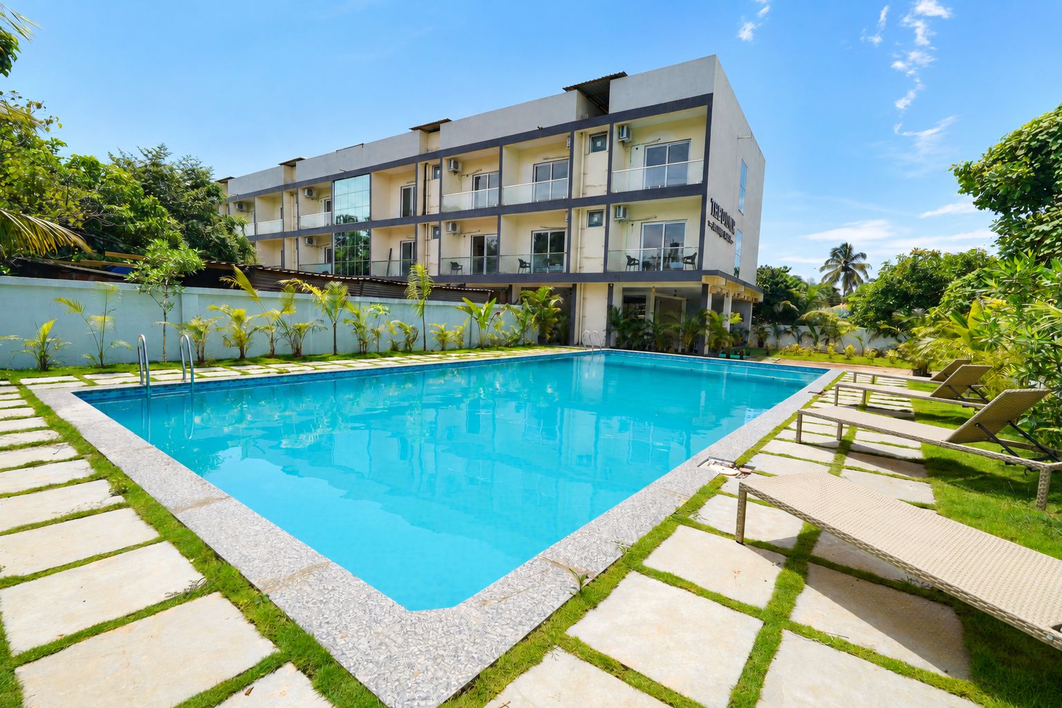 Ein modernes Hotel mit Schwimmbad, umgeben von einem gepflegten Garten und blauem Himmel.