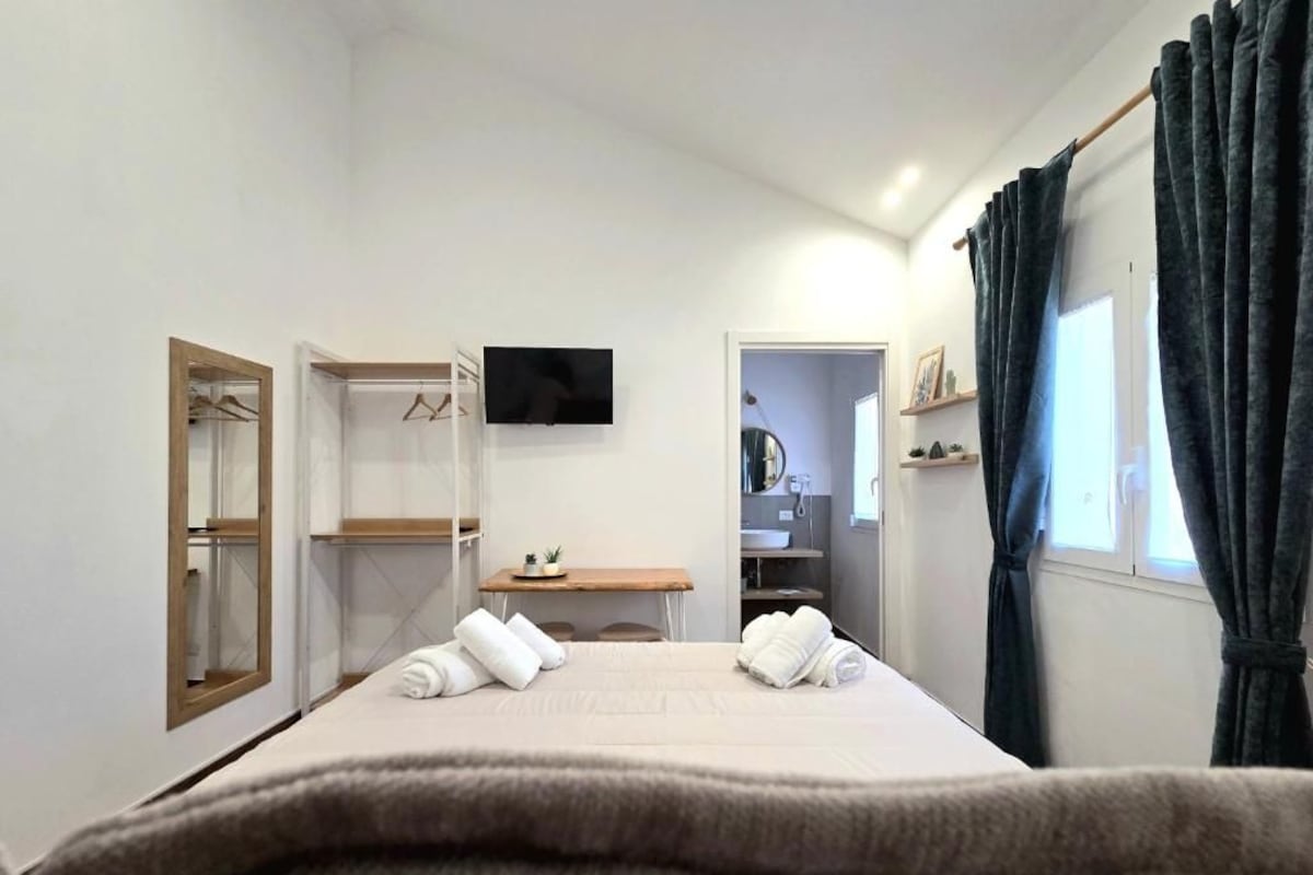 Hotellihuone, jossa on sänky, televisio, vaatekaappi, peili, työpöytä ja ikkuna, jossa on verhot.