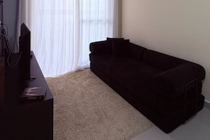 A room with a black sofa, a television on a stand, and a window with sheer curtains. - New Listing (Praia Grande)