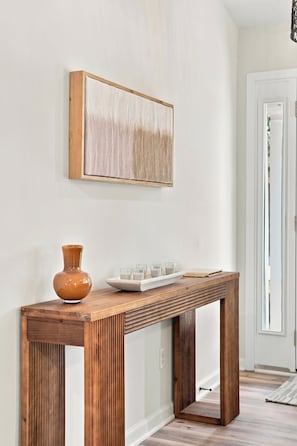 Une console en bois sur laquelle sont posés un vase et des verres, un tableau accroché au mur et une porte visible au fond.