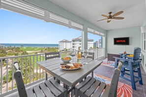 A balcony with a table set for a meal, a TV screen displaying 'NETFLIX', and a view of the ocean and buildings in the background. - Golf Views, Pool & Private Beach Access (Palm Coast)
