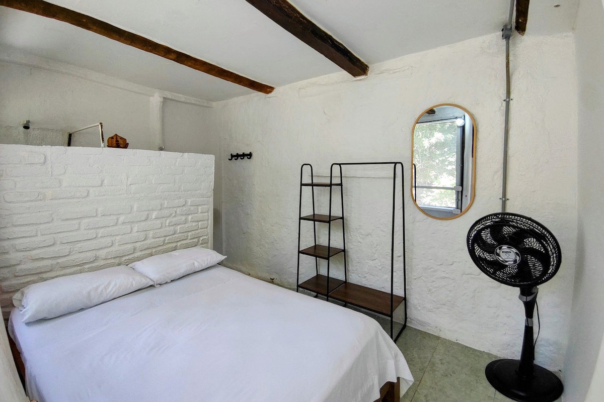 A room with a bed, a white brick wall, a metal shelf unit, a mirror, and a standing fan.