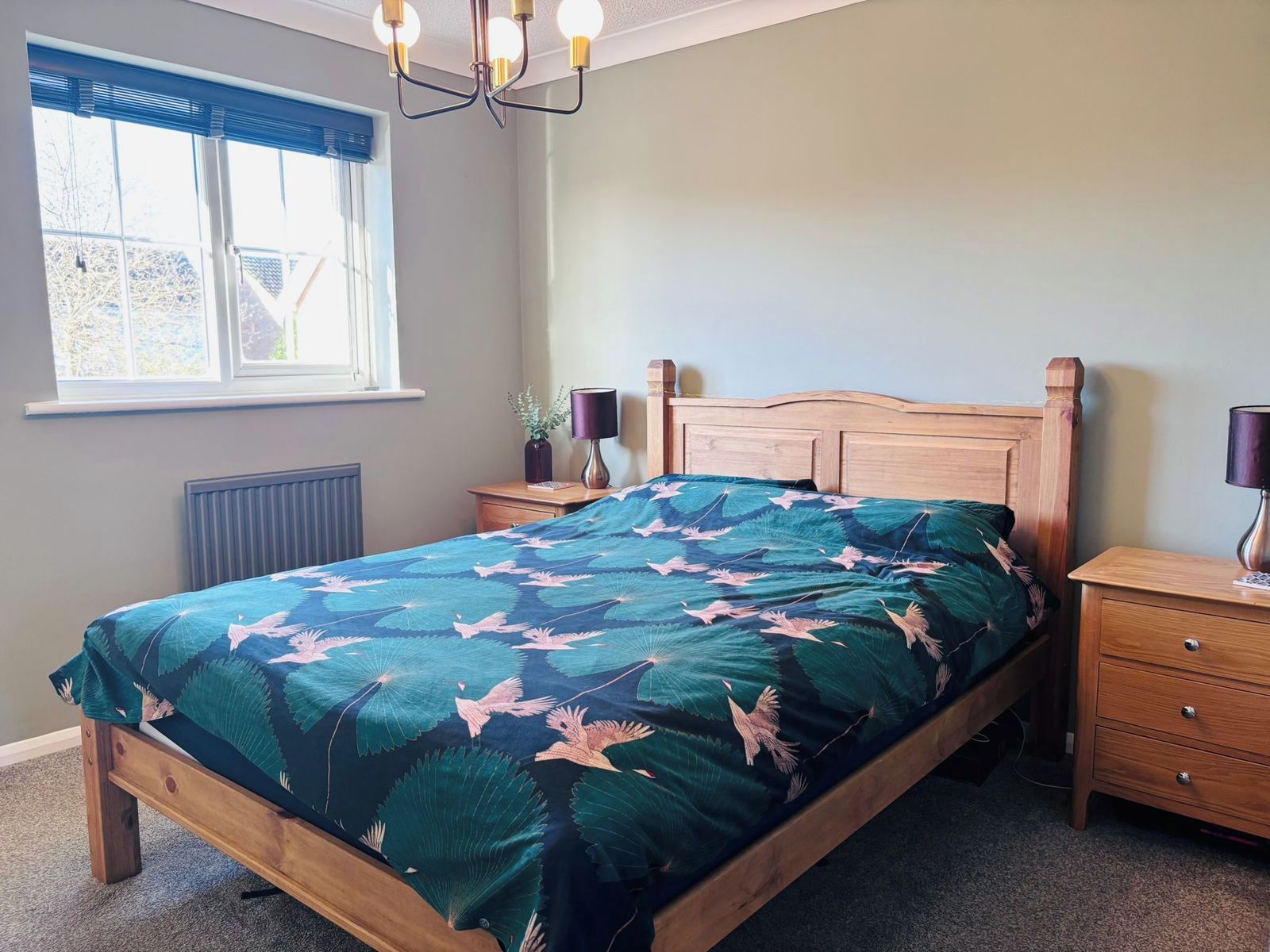 Une chambre à coucher avec un lit en bois, une table de chevet avec une lampe, une fenêtre avec des stores et un radiateur.