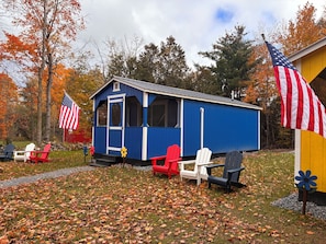 A small blue cabin with a door and windows, surrounded by autumn leaves and American flags.
