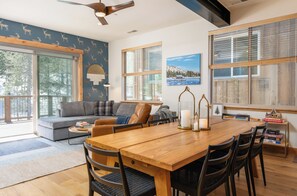 A modern living room with a wooden dining table, a sofa, and a large window with blinds. - Designer & Luxury Modern 3-Bed Penthouse w/Elevator in Truckee w/Hot Tub/Gym (Truckee)