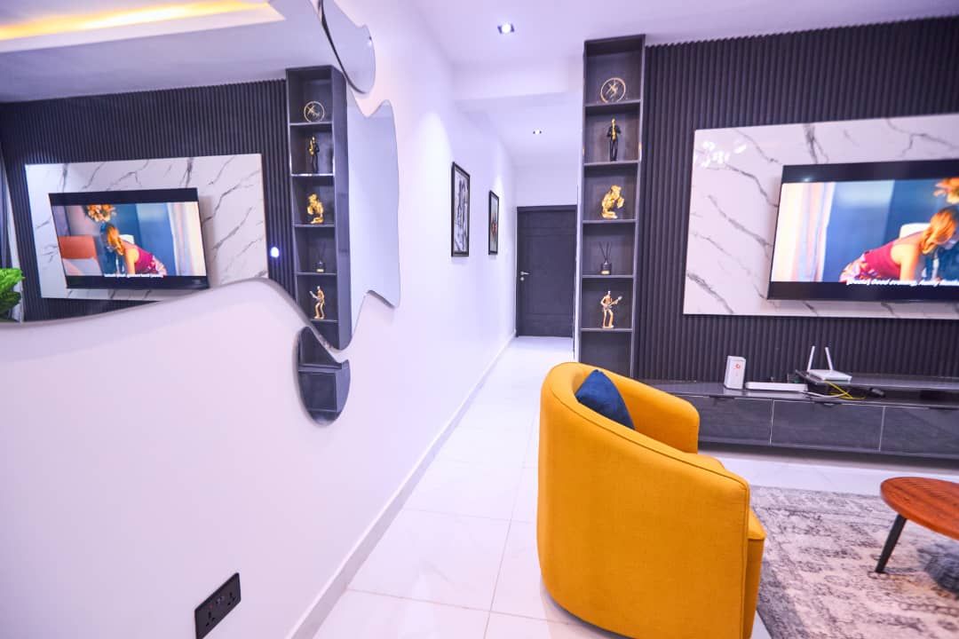 Una sala de estar moderna con dos televisores de pantalla plana grandes montados en la pared, un sillón amarillo y una mesa de centro de madera.