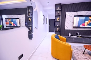 Ein modernes Wohnzimmer mit zwei großen Flachbildfernsehern an der Wand, einem gelben Sessel und einem Holztisch.