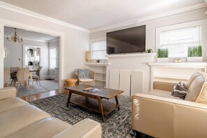 A modern living room with a flat-screen TV, a fireplace, and a dining area with a kitchen in the background. - A Block from Alberta St • 4BR • Sleeps 10 (Portland)