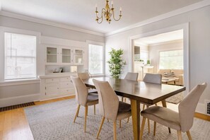 Een eetkamer met een grote tafel, stoelen, een kroonluchter en uitzicht op de aangrenzende woonkamer.