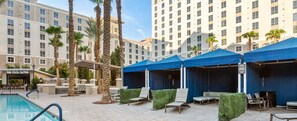 Un espace piscine agrémenté de chaises longues et de parasols, entouré de palmiers et d’un bâtiment d’hôtel.
