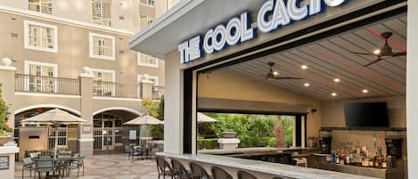 Un área de bar al aire libre con un letrero que dice 'THE COOL CAFE', mesas y sillas exteriores, y vistas del exterior del edificio.