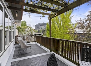 Ett takförsett utomhusområde med sittplatser, en pergola med ljusslingor och en utsikt över byggnader och träd i bakgrunden.