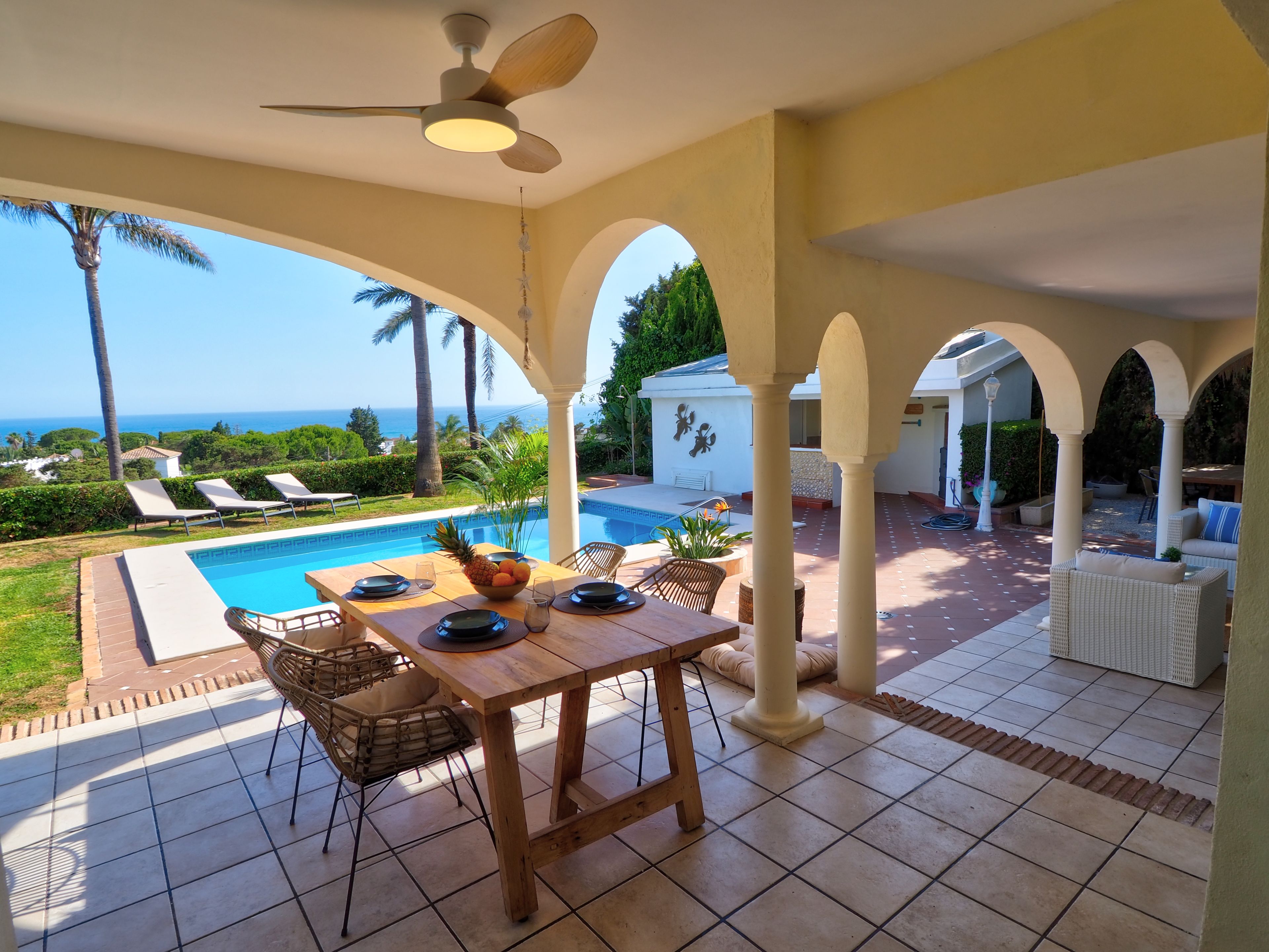 Ein überdachter Essbereich im Freien mit Pool, Liegestühlen und Blick aufs Meer.