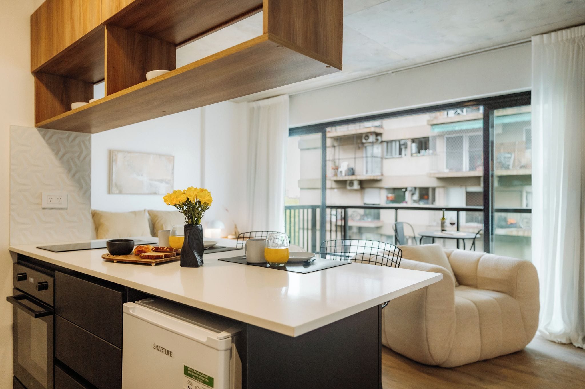 Een moderne keuken met een centraal eiland, een zithoek met een bank en uitzicht op een appartementencomplex door een glazen deur.