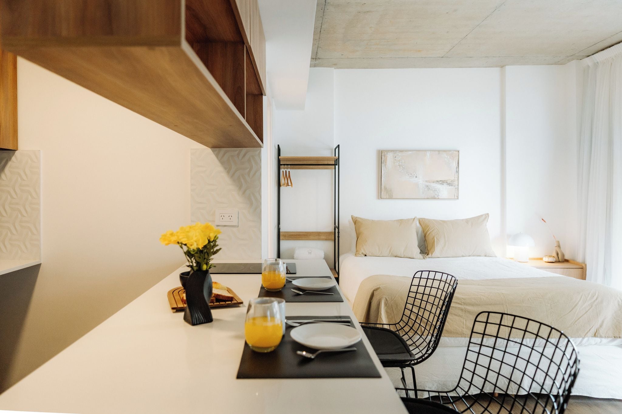 Een moderne hotelkamer met een zithoek, een bed met wit dekbedovertrek, een zwarte draadstoel en een wandrek.