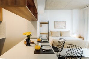 Een moderne hotelkamer met een zithoek, een bed met wit dekbedovertrek, een zwarte draadstoel en een wandrek.