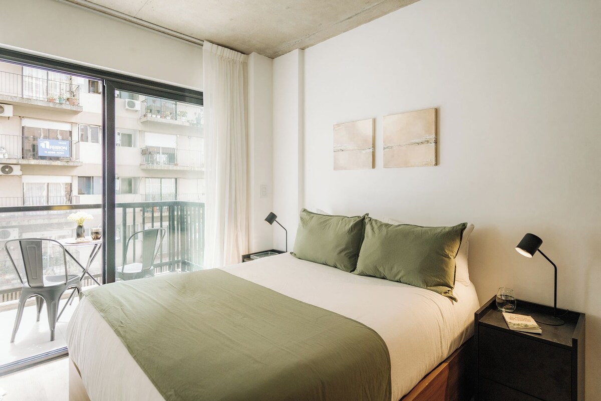 Een hotelkamer met een bed, twee nachtkastjes, een stoel, een balkon met uitzicht op gebouwen, en een raam met gordijnen.