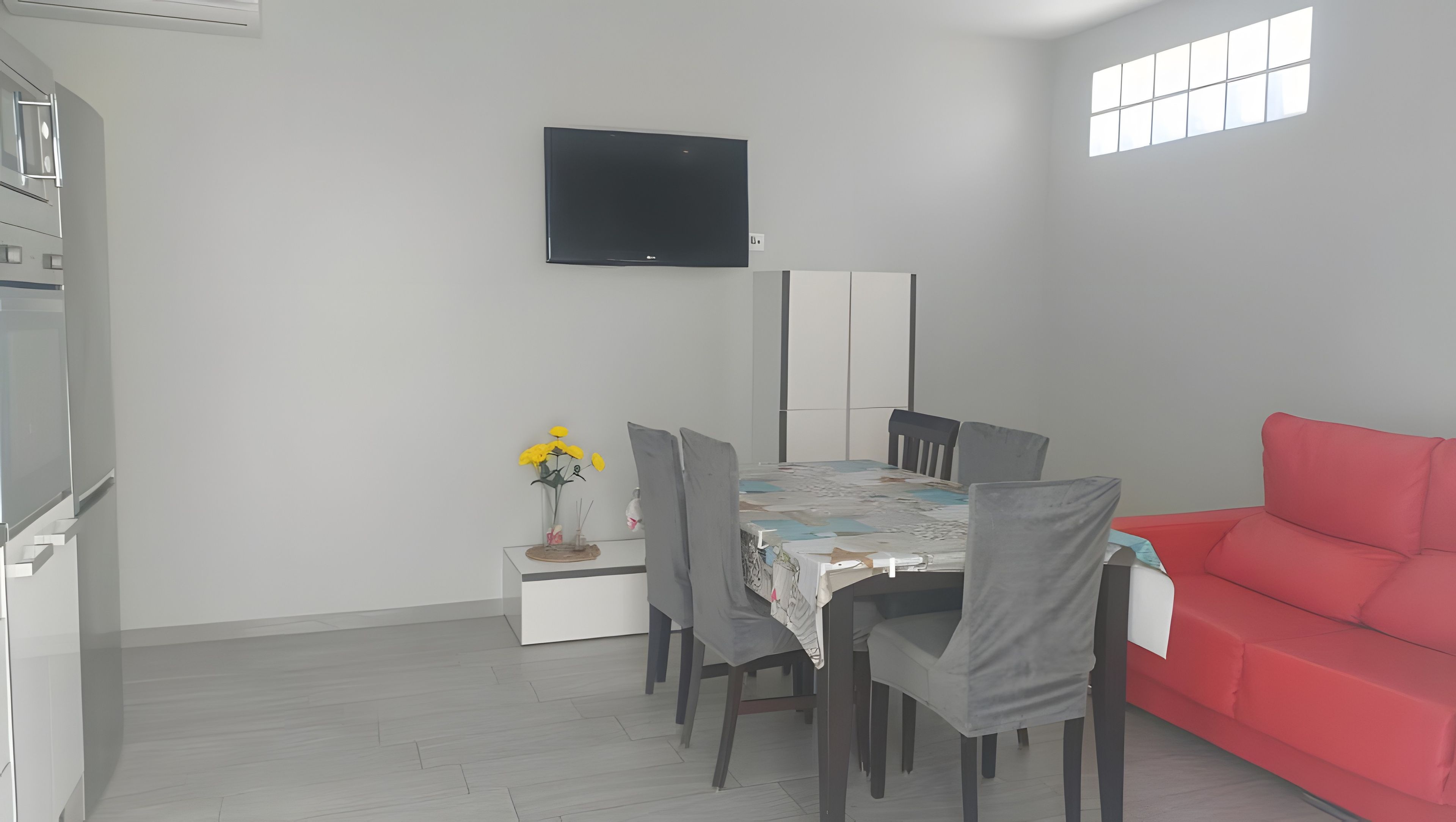 Ein modernes Zimmer mit Esstisch, Stühlen, einem roten Sofa, einem Flachbildfernseher und einer kleinen, dekorativen Vase mit Blumen.