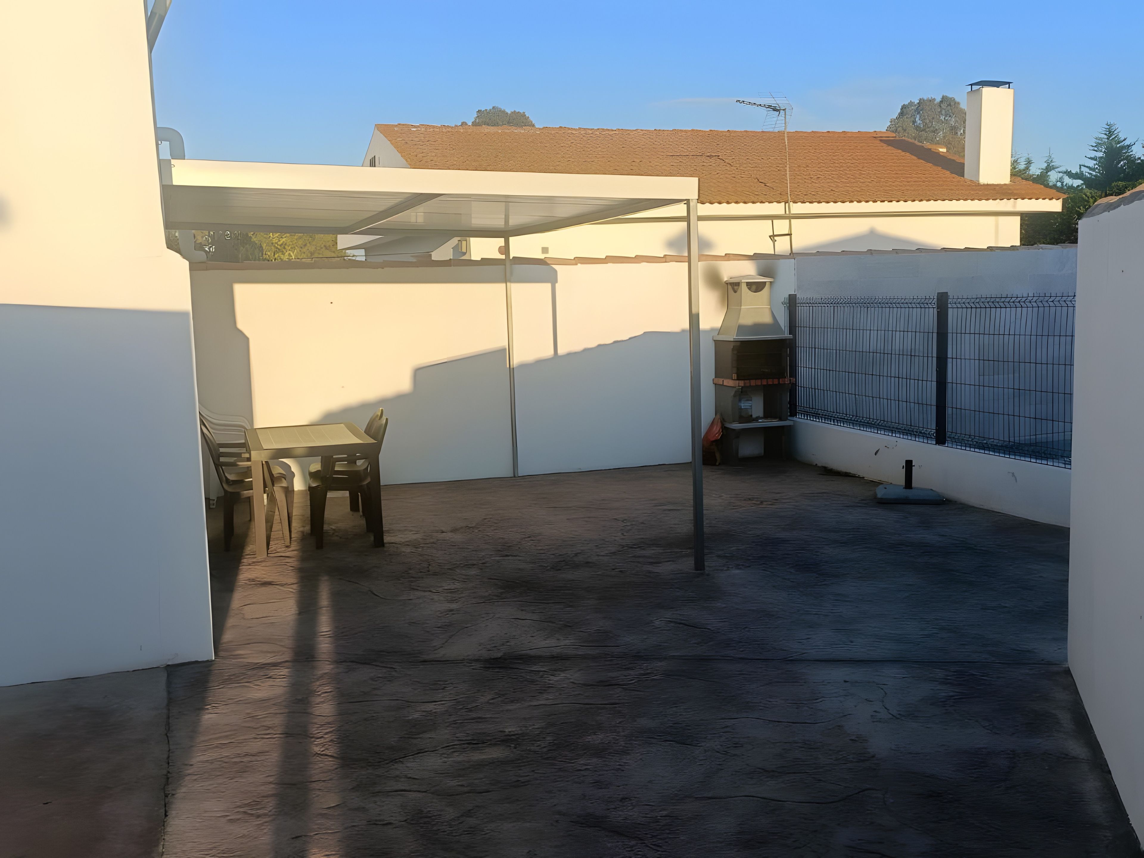 Een overdekt terras buiten, met een tafel, stoelen, een barbecue en een omheind gebied.