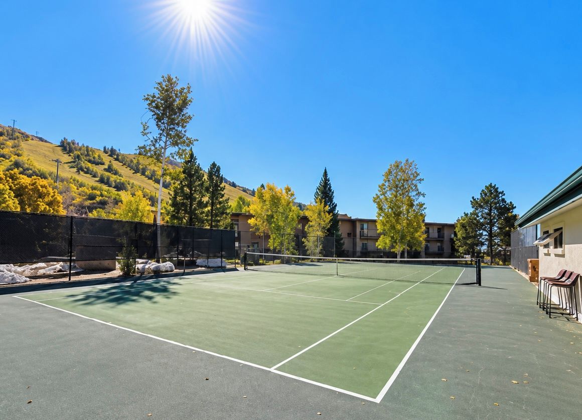 Una cancha de tenis exterior con red, rodeada de árboles y edificios.