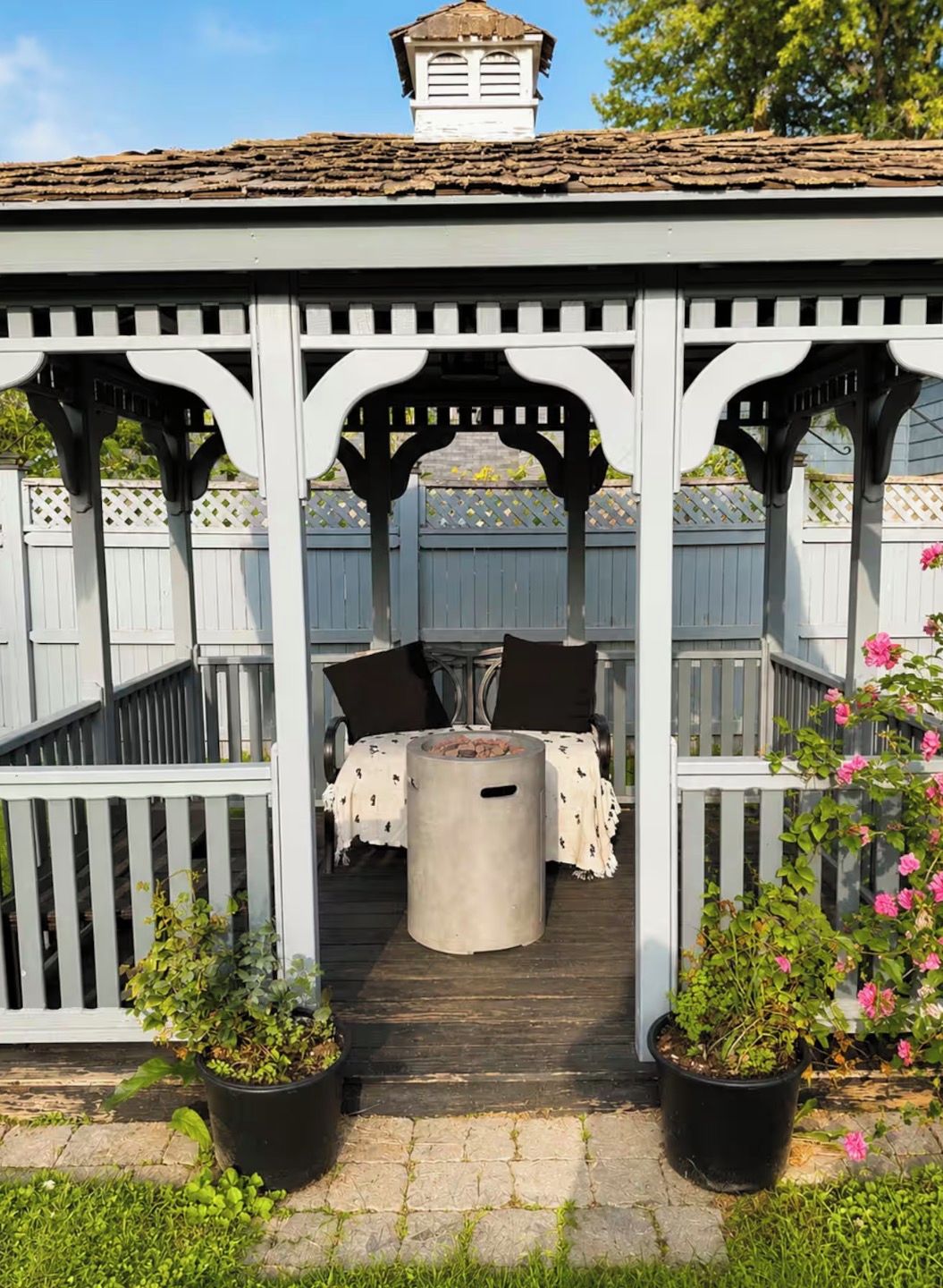 A covered outdoor seating area with a table and chairs, surrounded by a wooden fence and potted plants.