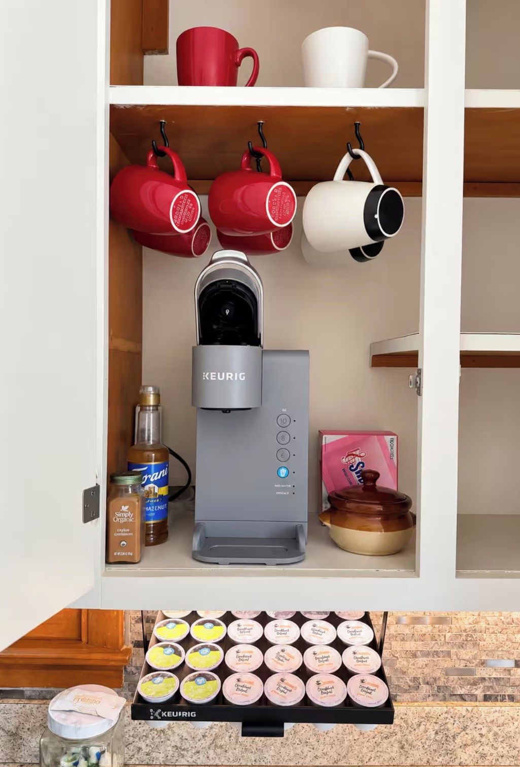 Et køkken med en Keurig kaffemaskine, røde og hvide krus, et glas med hasselnøddecreme, en sukkerdåse og en bakke med kaffekapsler.