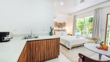 Una habitación de hotel moderna con kitchenette, una cama con ropa blanca, un comedor con mesa y sillas, y un ventanal con vistas a zonas verdes.
