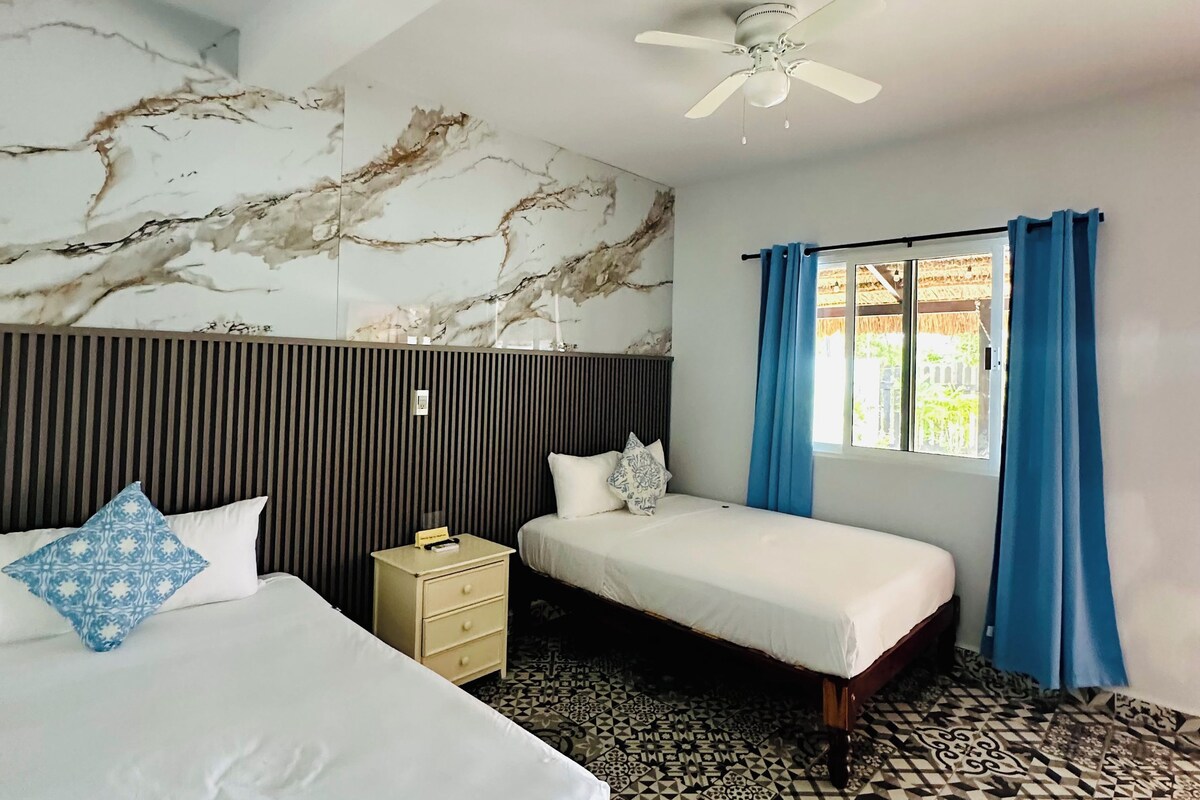 Une chambre d’hôtel avec un grand lit, un lit plus petit, une table de chevet, un ventilateur de plafond et des carreaux de sol à motifs.