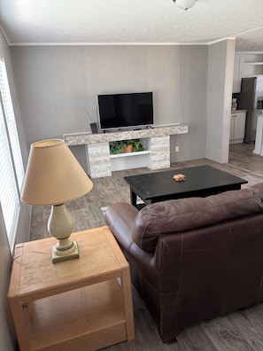 A living room with a television mounted above a fireplace, a brown leather sofa, a wooden side table with a lamp, and a black coffee table. - The Canal House on the canal with stunning sunsets. (Okeechobee)