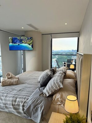 A modern bedroom with a large bed, a television mounted on the wall, and a view of a cityscape through a large window. - TERRA COTA | Stylish 3BR | Americana (Guadalajara)