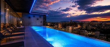 An infinity pool with a cityscape view, lounge chairs, and a modern building structure.