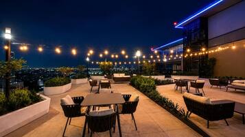 An outdoor seating area with tables and chairs, string lights, and a city view at night.