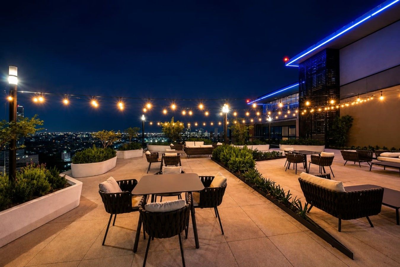 An outdoor seating area with tables and chairs, string lights, and a city view at night.