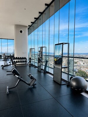 A gym with exercise equipment and large windows overlooking a cityscape. - CUR-CUMA | Boutique 2BR | Americana (Guadalajara)