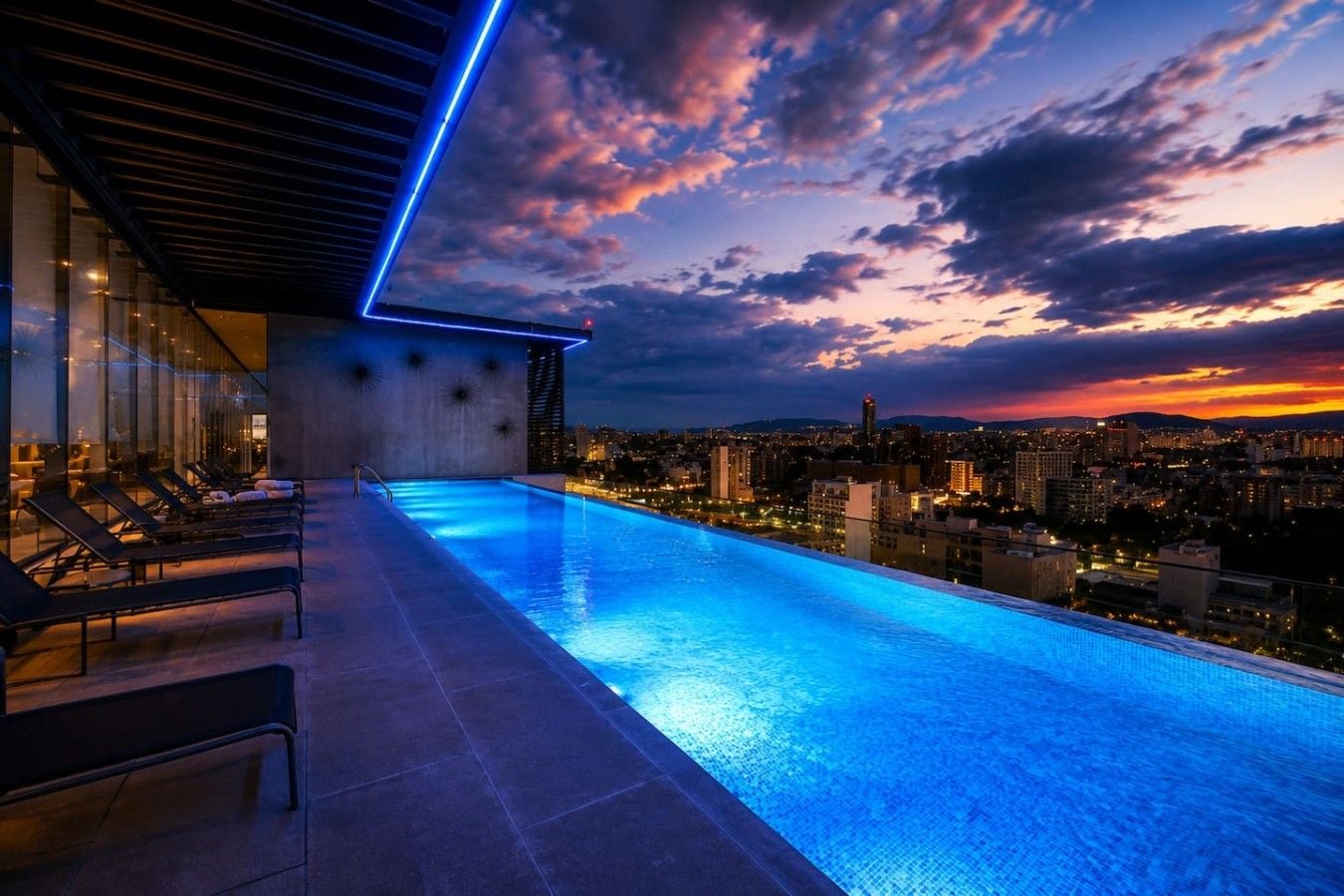 An infinity pool with a cityscape view, lounge chairs, and a modern building structure.