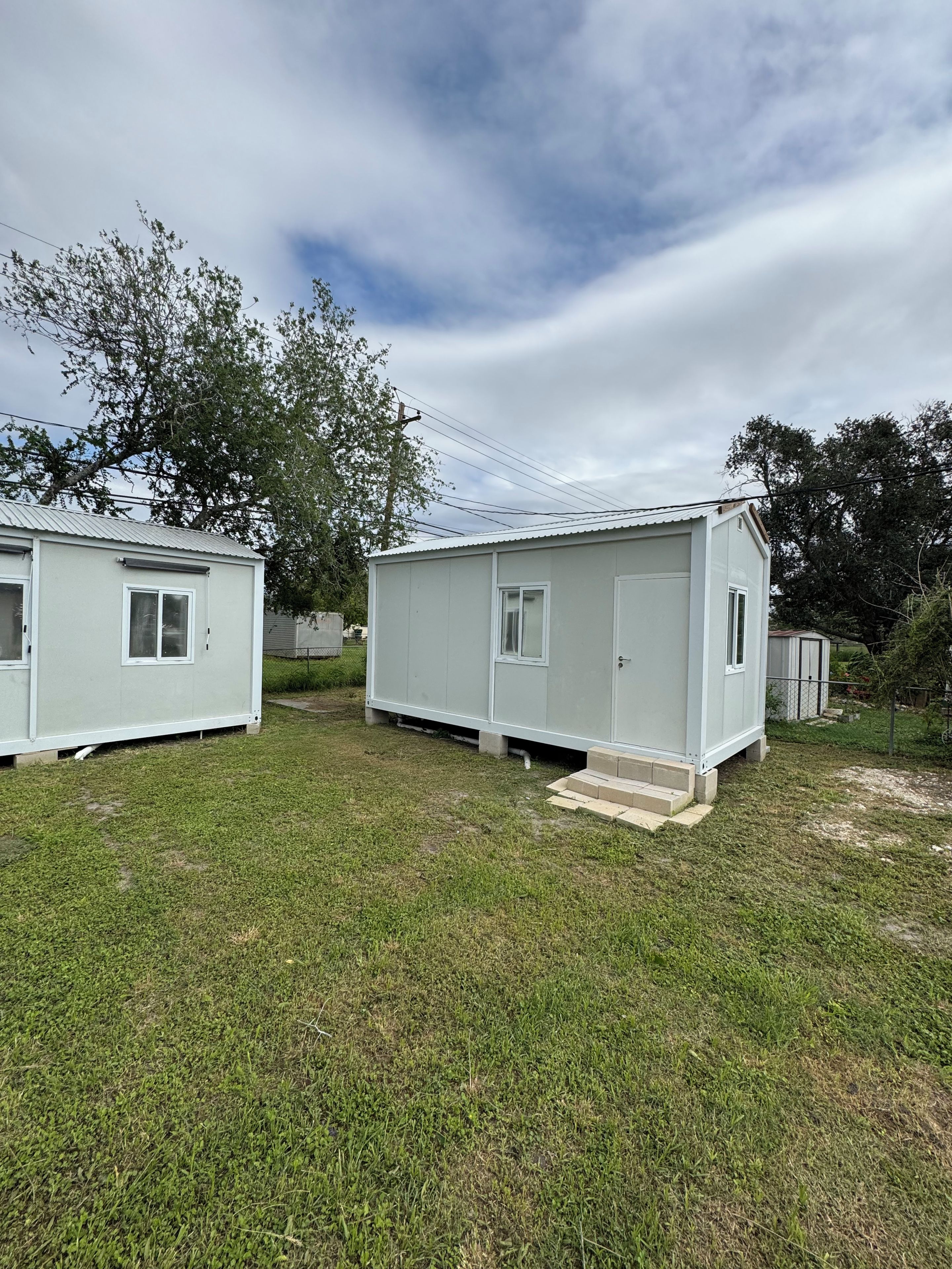 Two small, single-story mobile homes with a flat roof, a front door, and a small window on each side. They are situated on a grassy area with a tree in the background.