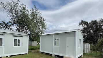 Two small, single-story mobile homes with a flat roof, a front door, and a small window on each side. They are situated on a grassy area with a tree in the background.