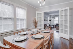 Comedor con mesa de madera dispuesta para cuatro personas, rodeada de sillas. Se observa un ventanal con persianas, un jarrón con plantas secas y una araña.