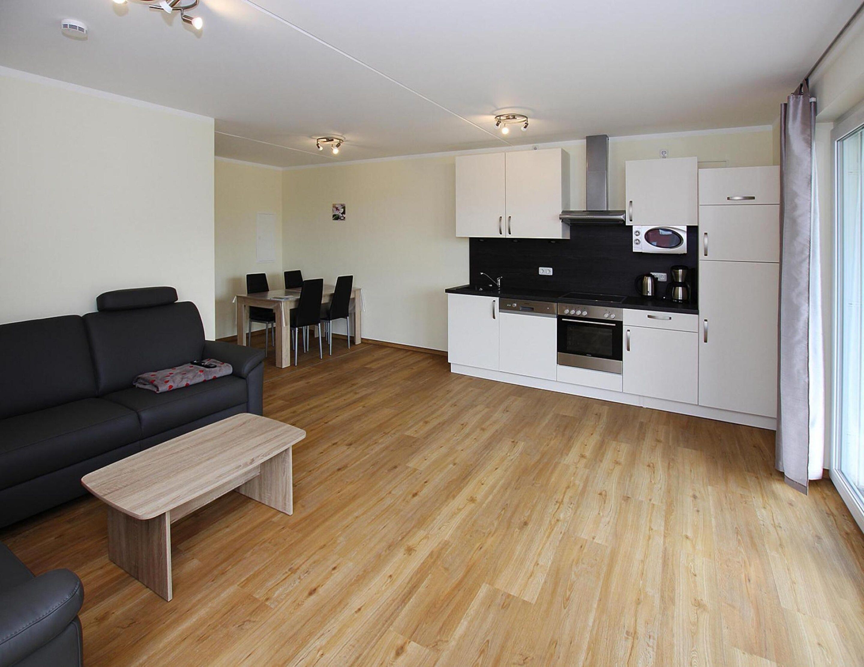 Ein modernes Wohnzimmer mit einem schwarzen Sofa, einem Holztisch und einem Essbereich mit Tisch und Stühlen. Eine Küche mit weißen Schränken und Geräten ist ebenfalls sichtbar.