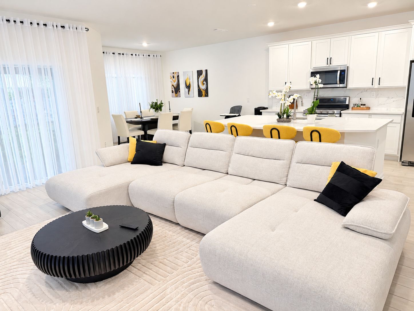 Un salon moderne avec un canapé d’angle, une table basse noire et un coin cuisine doté de meubles blancs et d’appareils électroménagers.