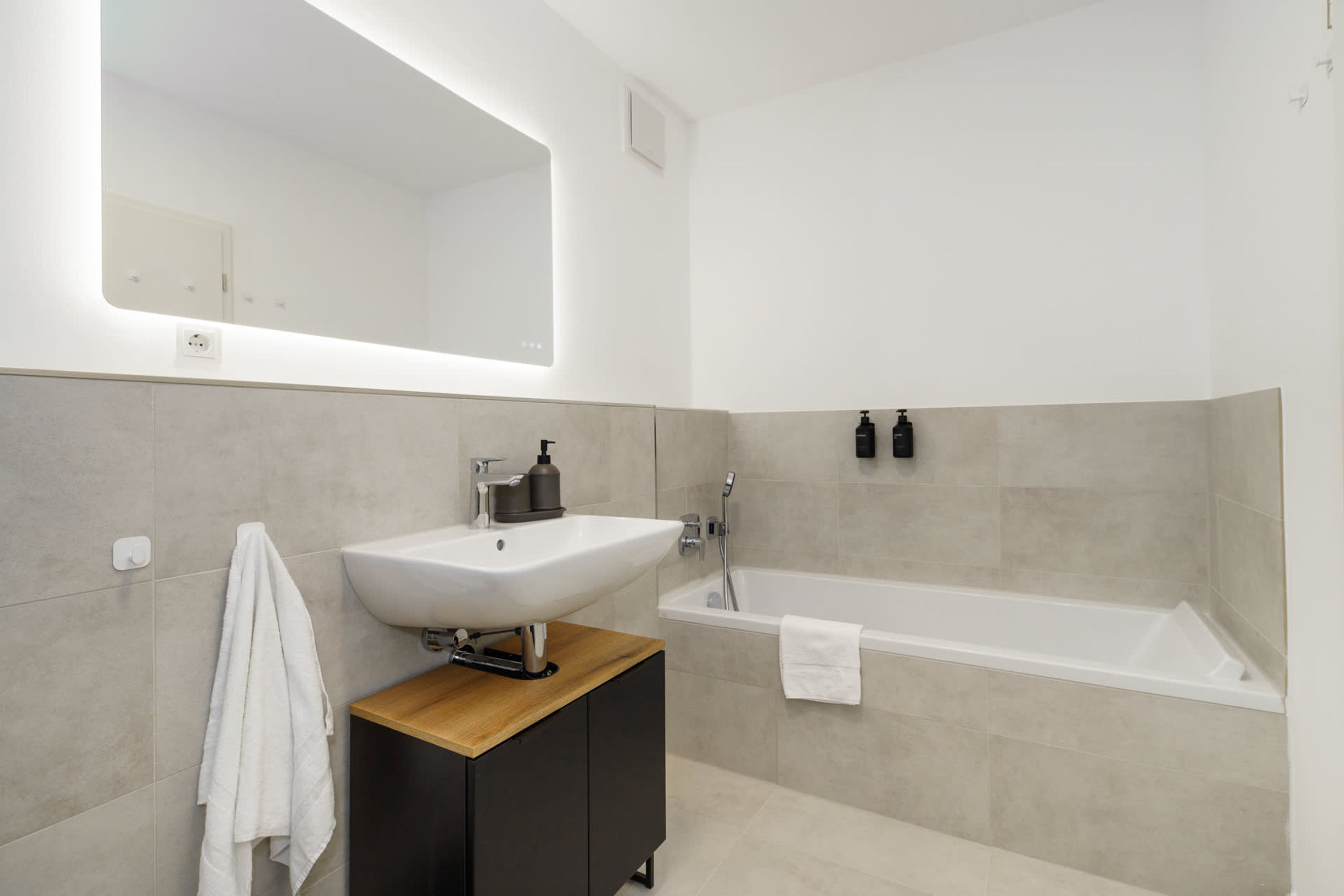 A modern bathroom with a white sink, a wooden vanity, a large mirror, and a bathtub.
