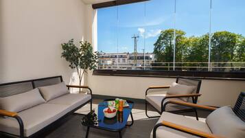 A balcony with a sofa, chairs, a table with a bowl of fruit and drinks, and a view of buildings and trees.