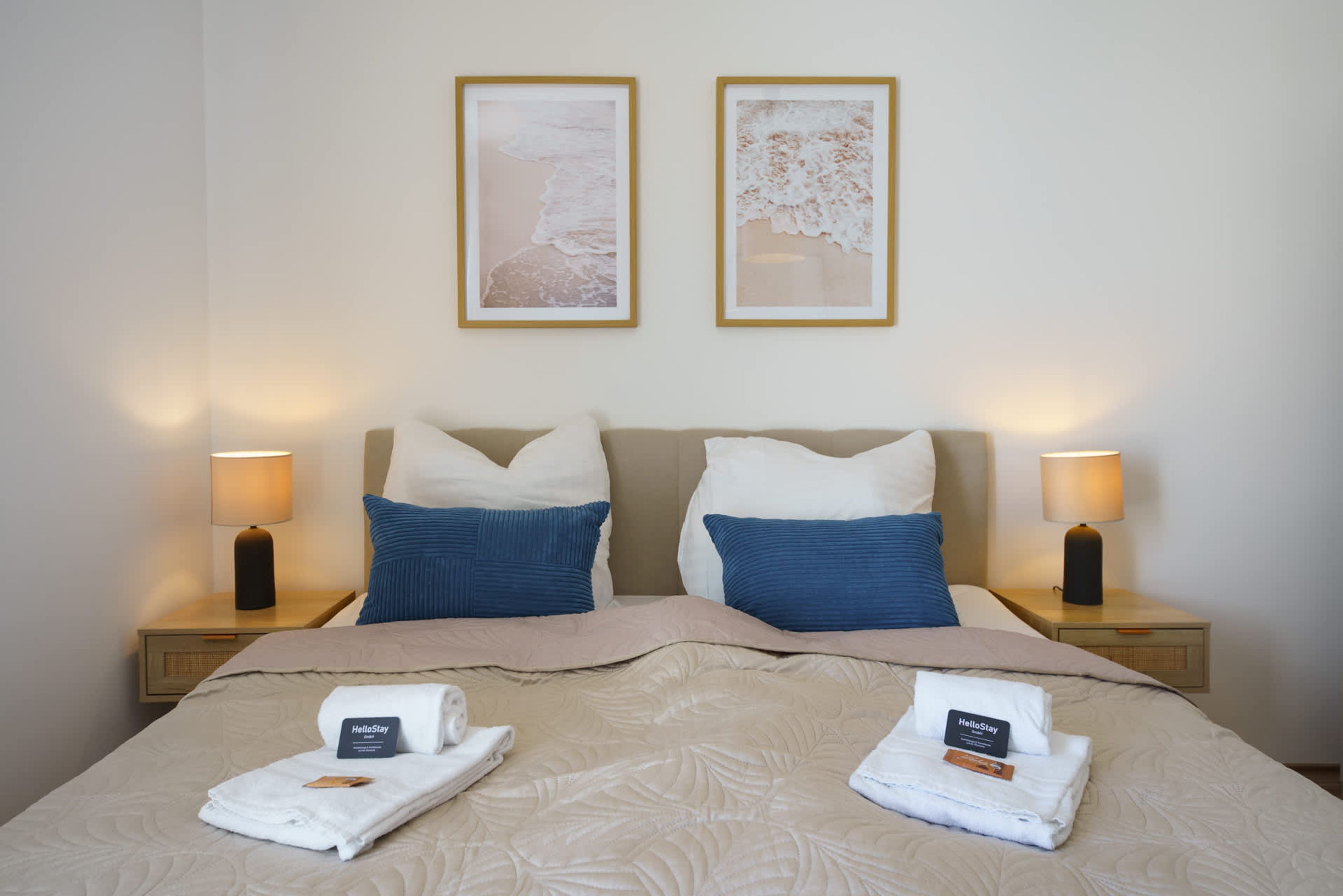 A hotel room with a bed, two pillows, two nightstands, two lamps, and two framed pictures on the wall.
