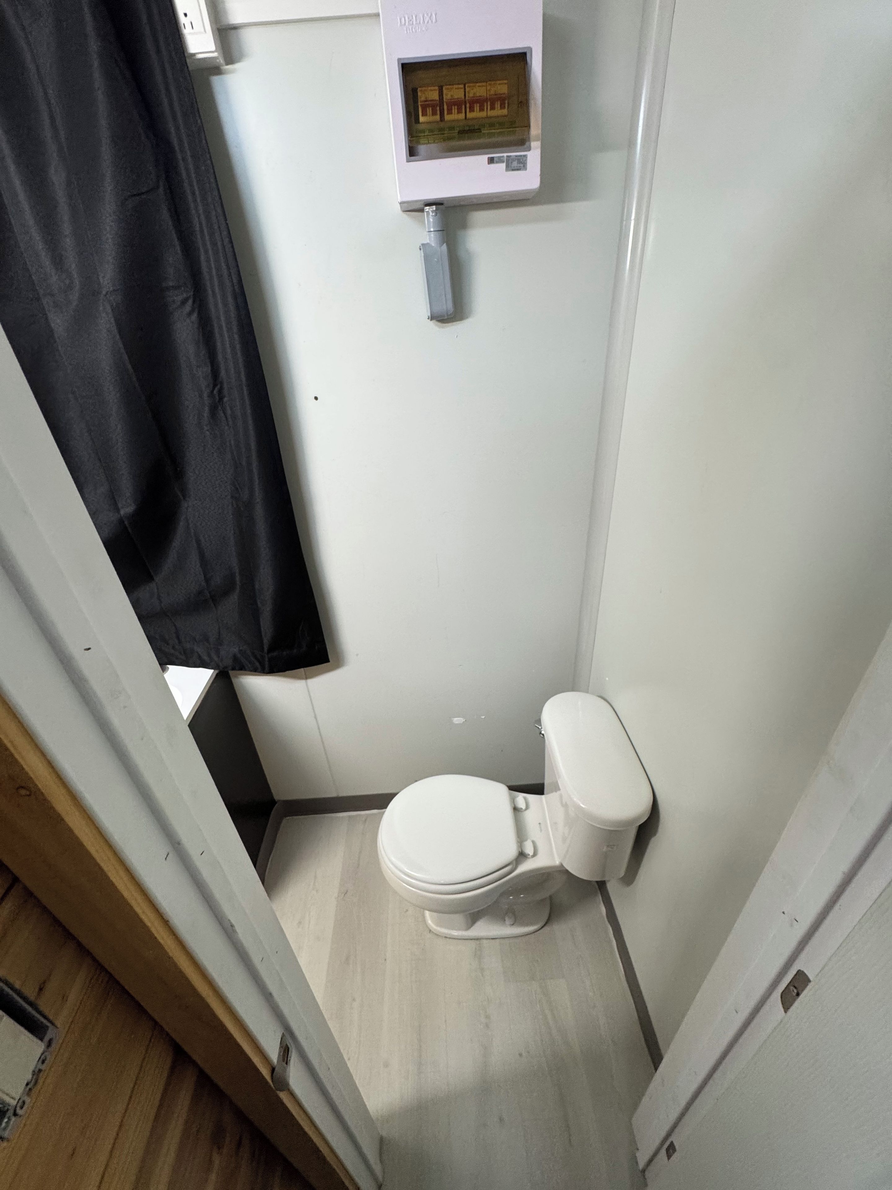 A small, compact bathroom with a toilet, a wall-mounted control panel, and a black shower curtain.