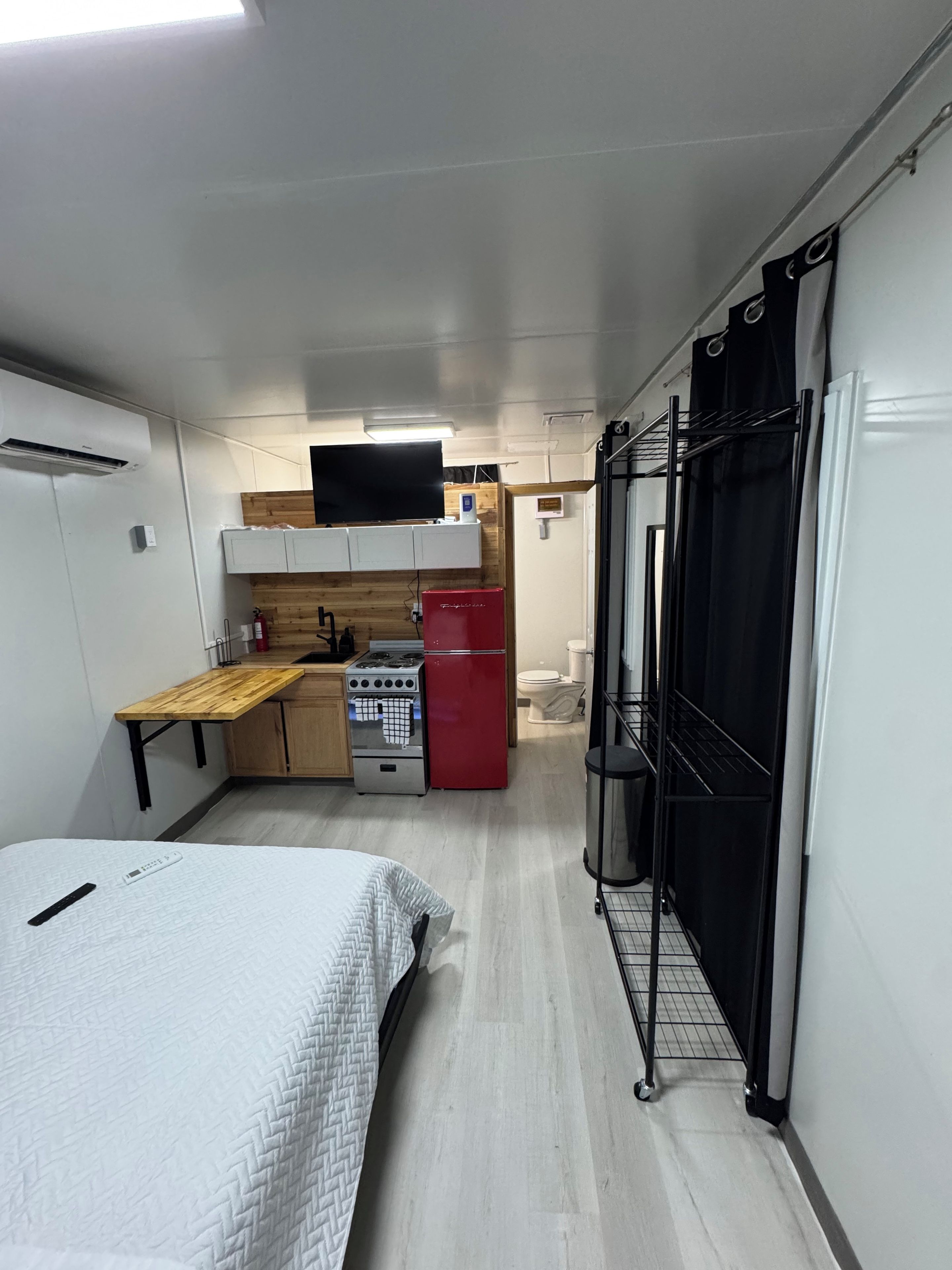 A compact room with a bed, a kitchenette with a stove, a refrigerator, and a small window.
