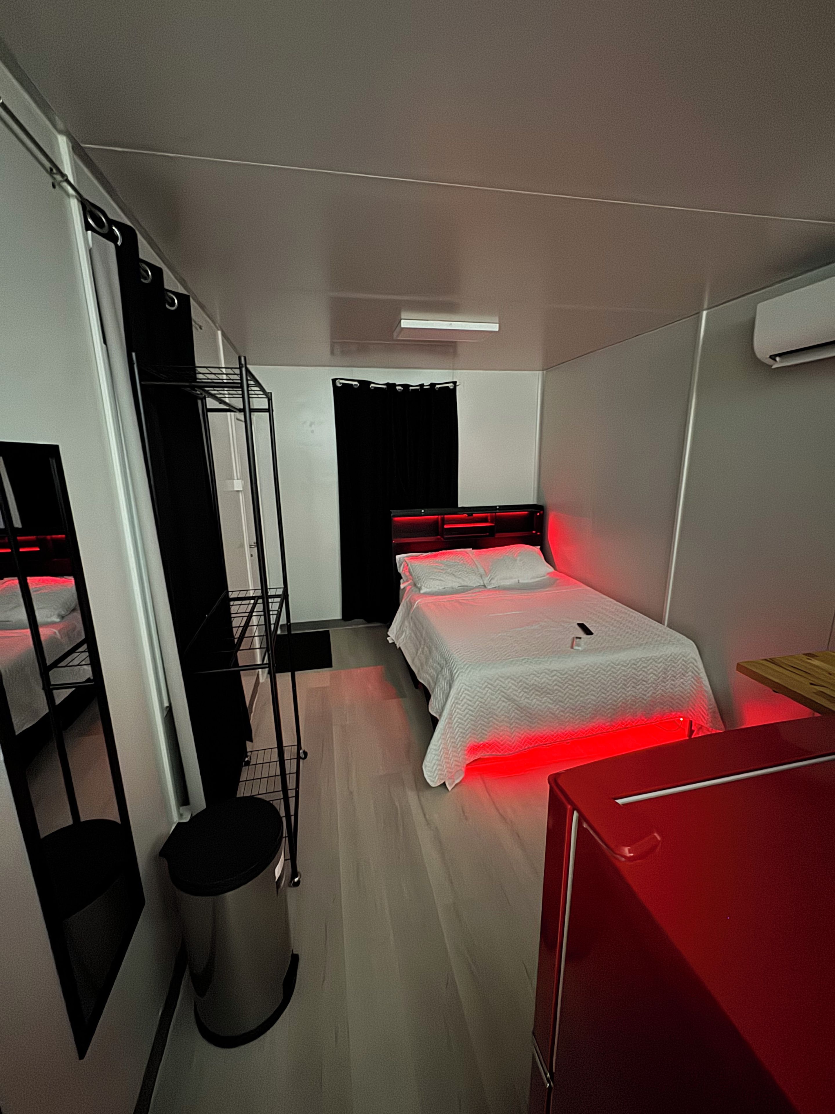 A compact hotel room with a bunk bed, a single bed with red lighting, a small trash bin, and a red desk.