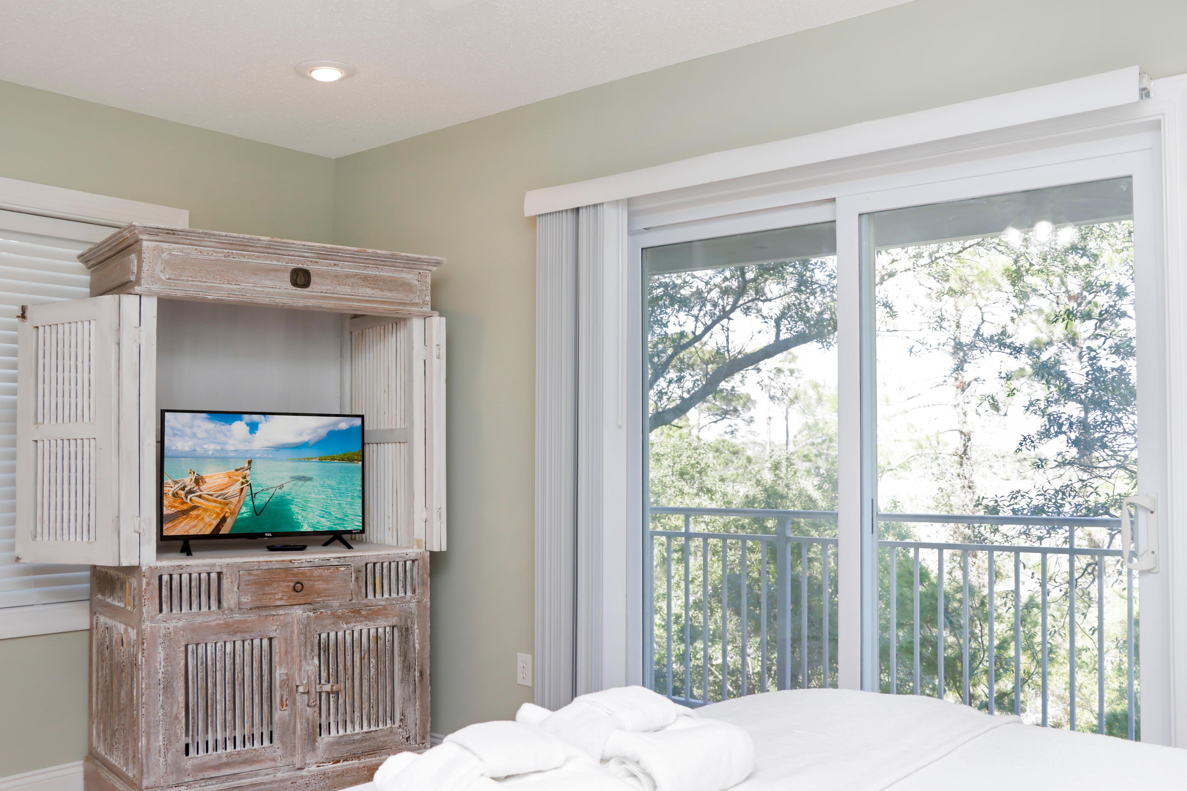 A bedroom with a television on a rustic wooden cabinet, a balcony with a railing, and a bed with white linens.