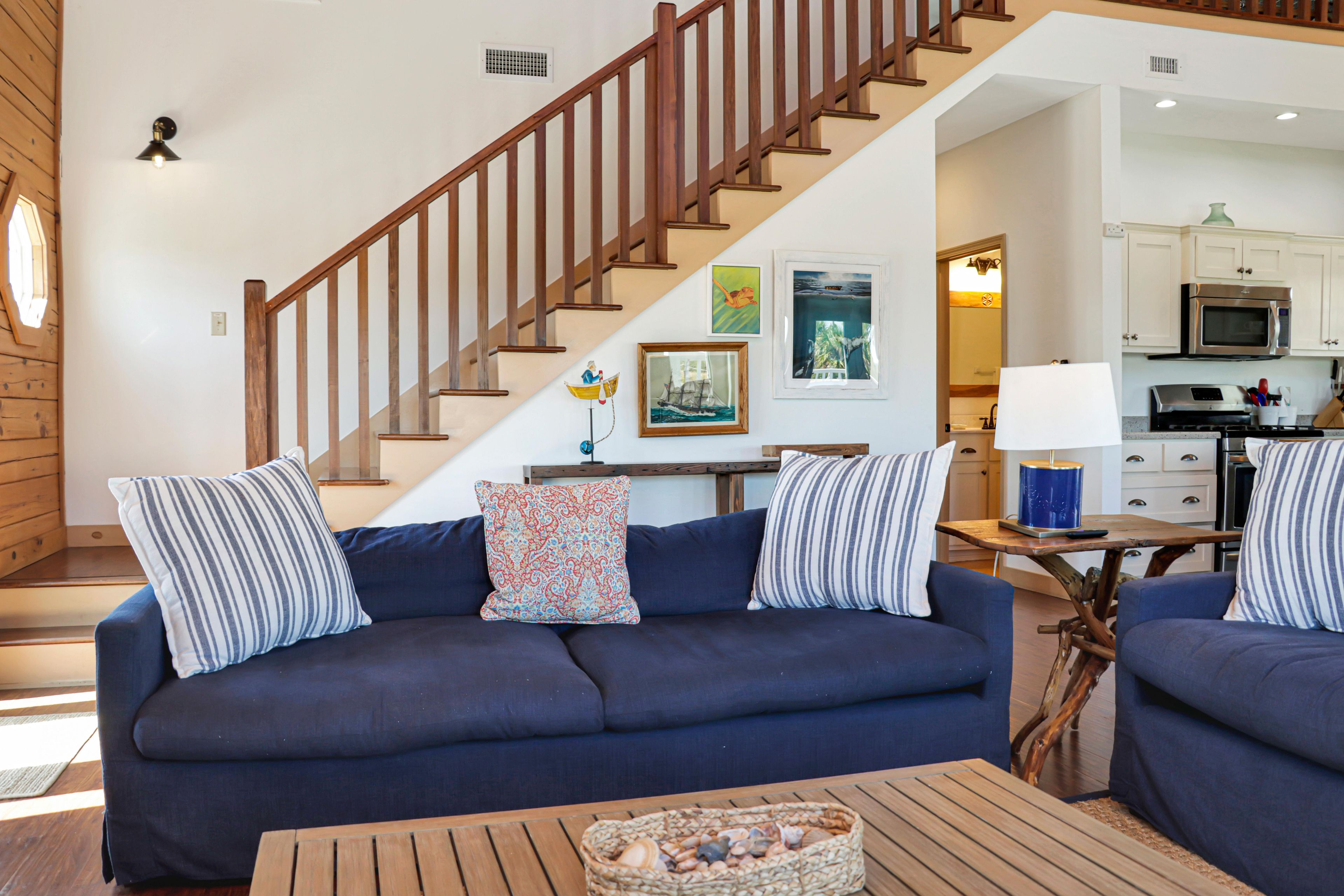 A living room with a dark blue sofa, wooden staircase, and a kitchen area in the background.