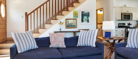 A living room with a dark blue sofa, wooden staircase, and a kitchen area in the background.